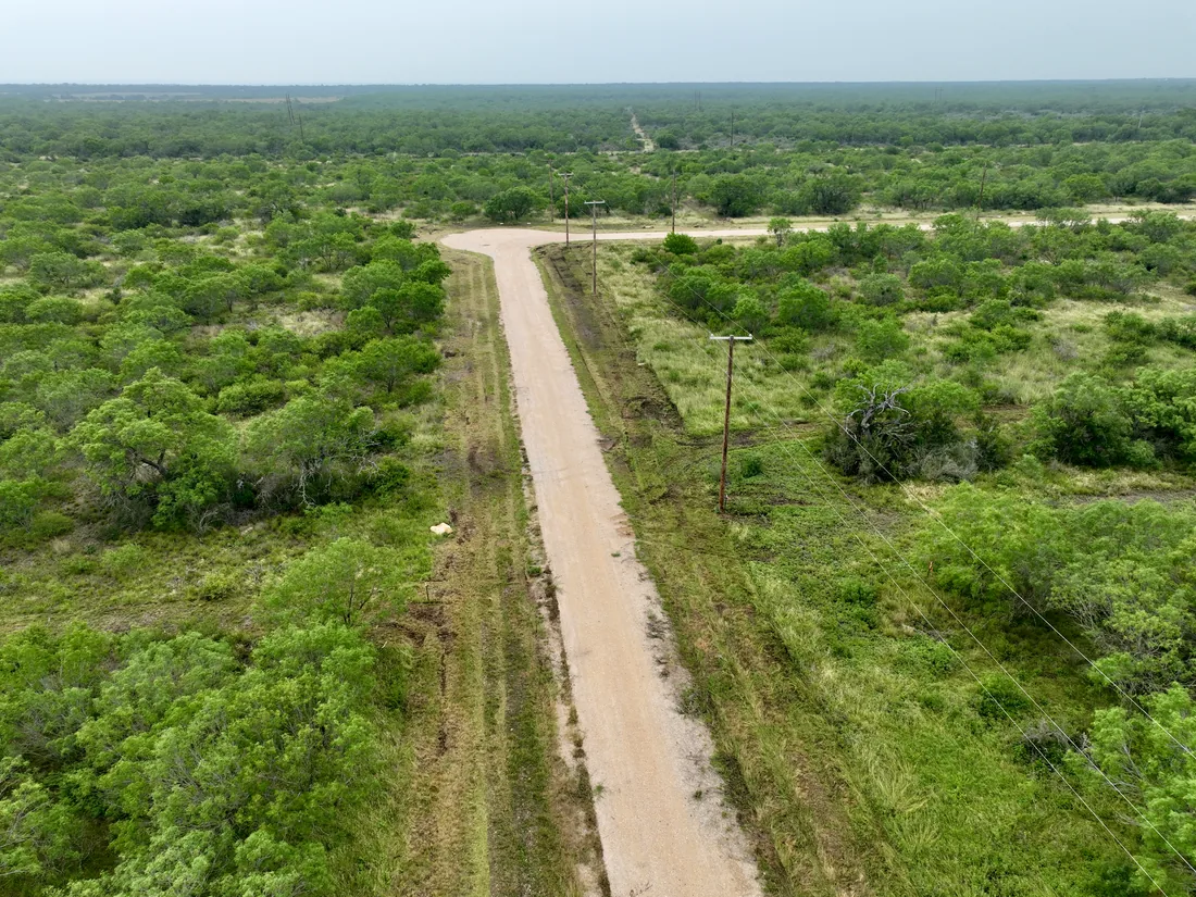 Low drone view down a maintained caliche road through green brush, with a pole line alongside